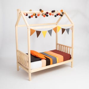 Natural solid wood toddler house bed frame on legs with colorful garland lights on a clean white backdrop.