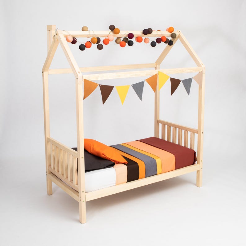 Natural solid wood toddler house bed frame on legs with colorful garland lights on a clean white backdrop.