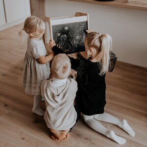 Three toddlers sitting on the floor drawing with chalk on the blackboard back panel of a wooden child standing stool.