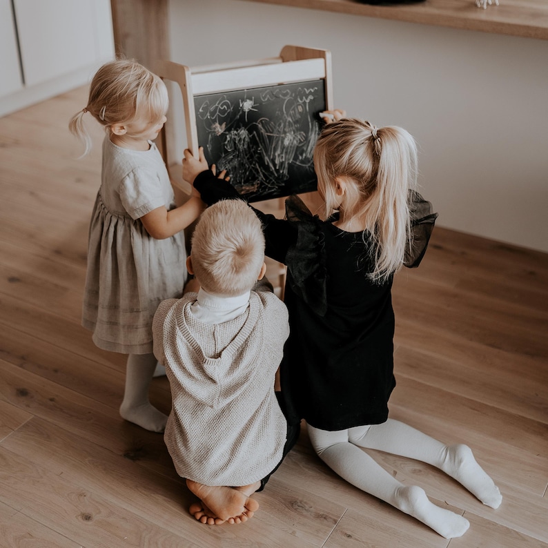 Three toddlers sitting on the floor drawing with chalk on the blackboard back panel of a wooden child standing stool.