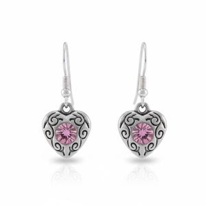May include: A pair of silver-toned heart-shaped earrings. Each earring features a pink gemstone at the center, surrounded by an ornate, swirling design. The earrings have a simple hook closure.