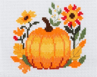 Può includere: Un motivo a punto croce su tela Aida bianca raffigurante una zucca arancione centrale circondata da fiori gialli e arancioni, foglie verdi e fogliame autunnale. Il disegno utilizza un motivo a punti a griglia per creare l'immagine.