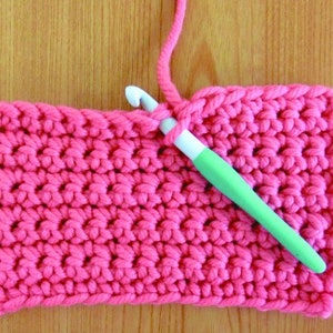 May include: A close-up of a pink crocheted fabric with a green crochet hook. The hook is being used to create a new stitch in the fabric.
