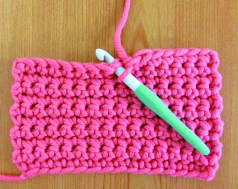 May include: A close-up of a pink crocheted fabric with a green crochet hook. The hook is being used to create a new stitch in the fabric.