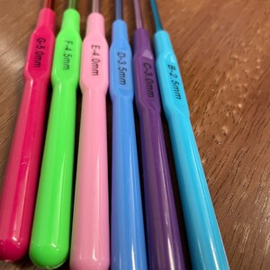 Pu&ograve; includere: Un set di sei ferri da uncinetto colorati con dimensioni etichettate da 2,5 mm a 5,0 mm. I ferri sono disposti in un motivo arcobaleno dal blu al rosa.