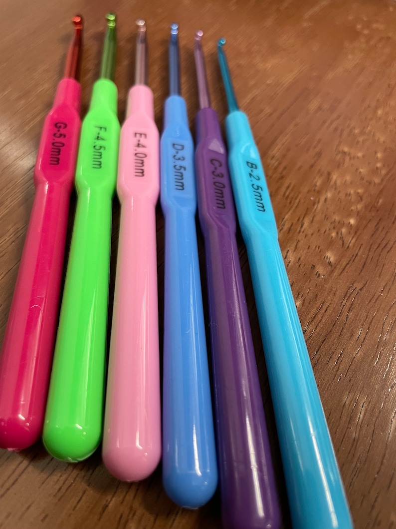 Pu&ograve; includere: Un set di sei ferri da uncinetto colorati con dimensioni etichettate da 2,5 mm a 5,0 mm. I ferri sono disposti in un motivo arcobaleno dal blu al rosa.