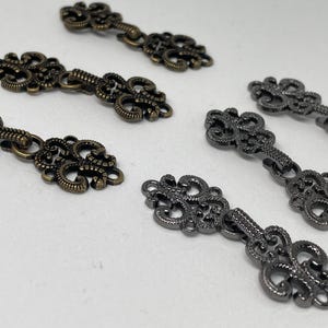 Puede incluir: Dos juegos de cierres metálicos decorativos, un juego en latón antiguo y un juego en negro. Cada cierre tiene dos piezas ornamentadas conectadas por una cadena.