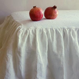 May include: Two ripe pomegranates rest on a white, ruffled tablecloth. The table covering has a soft, textured appearance, with a gathered edge that adds a decorative element. The pomegranates are a deep red color.