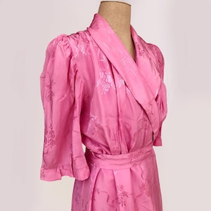 May include: A vibrant pink vintage dressing gown with a floral design. It has a shawl collar, three-quarter sleeves with frilled cuffs, and a matching belt. The fabric has a subtle sheen, and the gown is displayed on a mannequin.