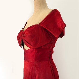 Può includere: Un abito rosso vibrante con un dettaglio a fiocco unico sul busto e uno scollo a cuore. L'abito presenta un corpetto aderente e una gonna fluida, mostrando una silhouette classica. Il tessuto ha una lucentezza sottile, che ne aumenta l'eleganza.