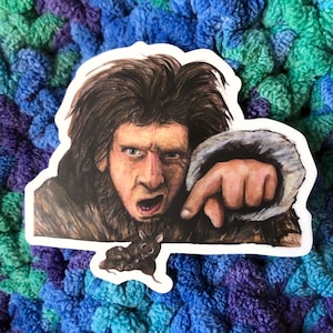 May include: A sticker featuring a painting of a man with long brown hair and a beard, wearing a brown fur coat. He is pointing with his right hand and has a surprised expression on his face. There are three small black rats in the bottom right corner of the sticker.
