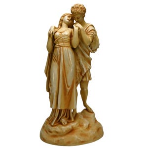 May include: A cream-colored ceramic sculpture of a couple embracing. The man is on the right and the woman is on the left. They are both standing on a rocky base.