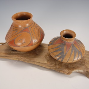 May include: Two small, hand-painted pottery vases with geometric designs in shades of brown, orange, and black. The vases are displayed on a piece of driftwood.