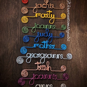 May include: A collection of ten custom wire names in various colors including red, copper, yellow, green, blue, light blue, silver, rose gold, magenta, and white. Each name features a spiral at both ends. The text Wirecreationsbyjules is visible.