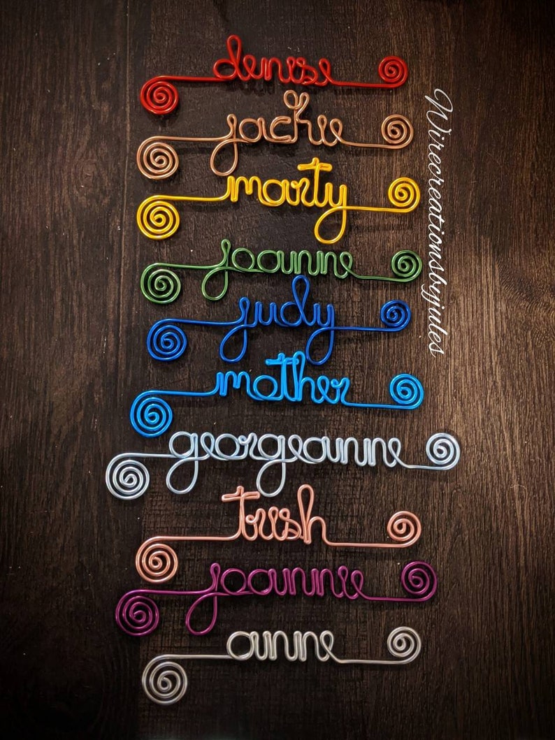 May include: A collection of ten custom wire names in various colors including red, copper, yellow, green, blue, light blue, silver, rose gold, magenta, and white. Each name features a spiral at both ends. The text Wirecreationsbyjules is visible.