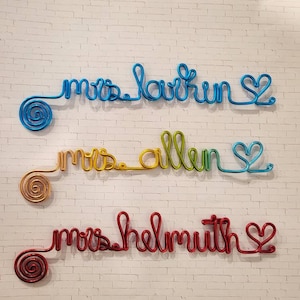 May include: Three custom wire wall signs featuring cursive text. The top sign is blue and reads "mrs larkin". The middle sign is a yellow-to-green gradient and reads "mrs allen". The bottom sign is red and reads "mrs helmuth". Each sign ends with a heart.
