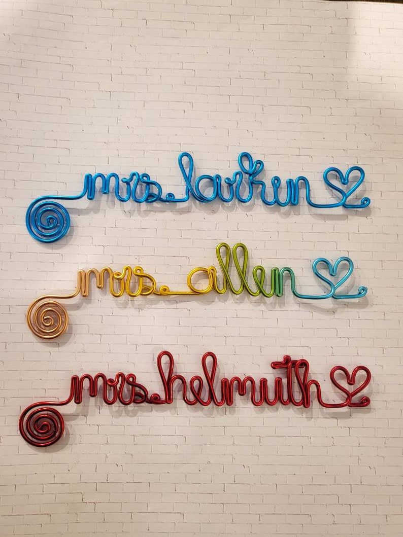 May include: Three custom wire wall signs featuring cursive text. The top sign is blue and reads "mrs larkin". The middle sign is a yellow-to-green gradient and reads "mrs allen". The bottom sign is red and reads "mrs helmuth". Each sign ends with a heart.