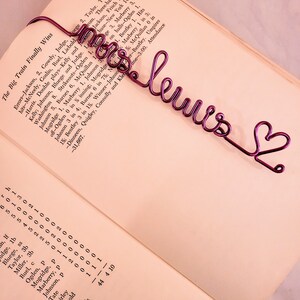 May include: A purple wire bookmark shaped into the cursive text "mrs lewis" followed by a small heart outline. The bookmark is placed on the open page of a book containing printed text and statistical data about a baseball game.