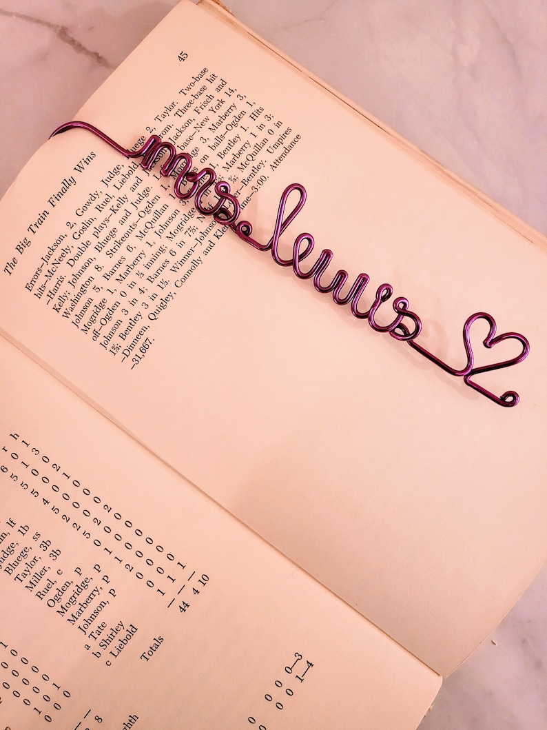 May include: A purple wire bookmark shaped into the cursive text "mrs lewis" followed by a small heart outline. The bookmark is placed on the open page of a book containing printed text and statistical data about a baseball game.