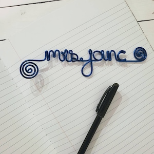 May include: A blue wire sculpture forming the cursive text "mrs. jonc" with a spiral design at each end, positioned on a lined notebook page next to a black ballpoint pen. The wire is a vibrant, glossy blue color and features a continuous script.