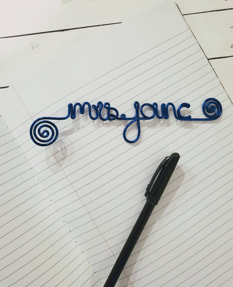May include: A blue wire sculpture forming the cursive text "mrs. jonc" with a spiral design at each end, positioned on a lined notebook page next to a black ballpoint pen. The wire is a vibrant, glossy blue color and features a continuous script.
