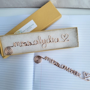 May include: Two copper-colored wire bookmarks, one resting inside a rectangular yellow gift box with a white label reading Wire Creations By Jules, and one lying flat. Each bookmark features a spiral end, cursive text, and a heart-shaped terminal.