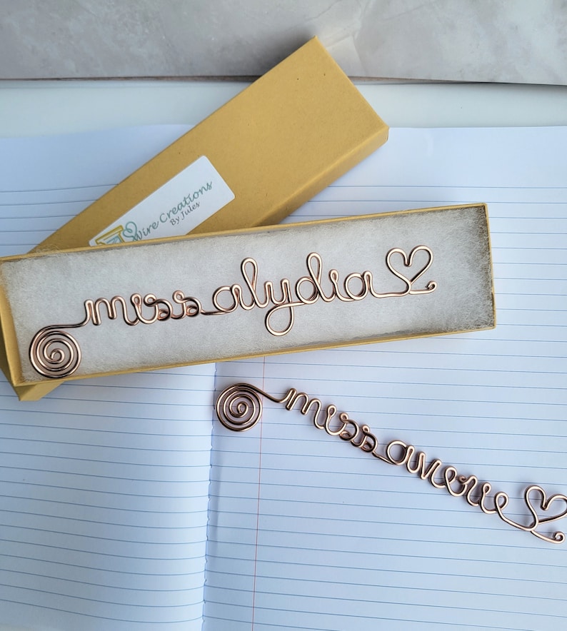 May include: Two copper-colored wire bookmarks, one resting inside a rectangular yellow gift box with a white label reading Wire Creations By Jules, and one lying flat. Each bookmark features a spiral end, cursive text, and a heart-shaped terminal.
