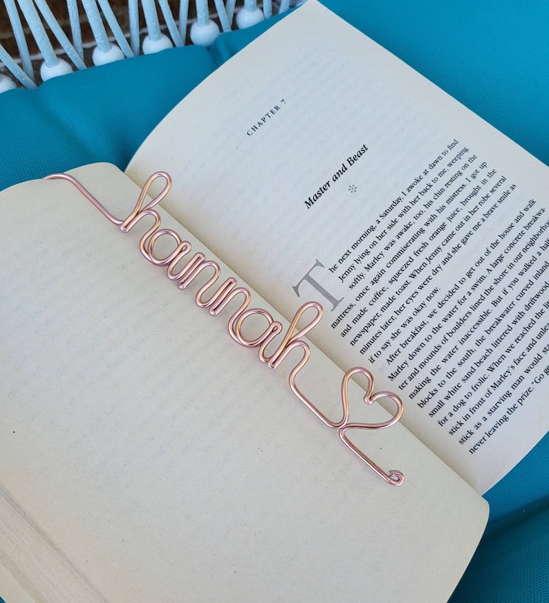 May include: A rose gold colored metal wire bookmark shaped into the cursive name Hannah, ending with a small heart outline. The bookmark is placed on the open pages of a book showing the text Chapter 7 Master and Beast on the white paper.