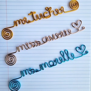 May include: Three custom wire name signs in gold, rose gold, and blue. The gold wire reads "mr tucker", the rose gold wire reads "miss avery", and the blue wire reads "ms morelle". Each wire sign features a spiral end and cursive lettering.
