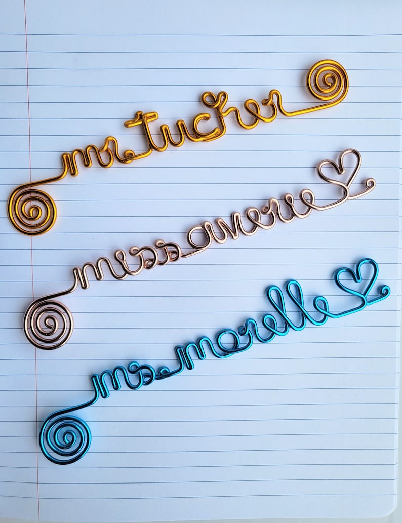 May include: Three custom wire name signs in gold, rose gold, and blue. The gold wire reads "mr tucker", the rose gold wire reads "miss avery", and the blue wire reads "ms morelle". Each wire sign features a spiral end and cursive lettering.