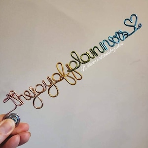 May include: A wire sculpture of the words "Thoughtful Planner" in gold, green, and blue wire. The sculpture is held in a hand. The words "Wirecreationsbyjules" are written in white text below the sculpture.