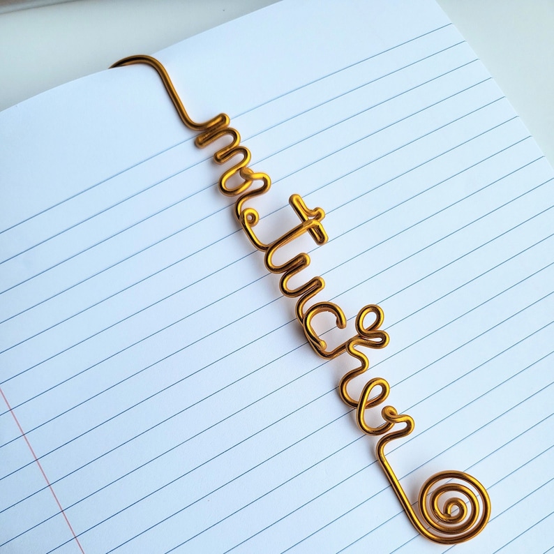 May include: A gold-colored metal wire bookmark shaped into the cursive word "mystic" with a spiral end. The bookmark is positioned diagonally across a white page with blue horizontal lines, showing the wire bent to hook over the top edge.
