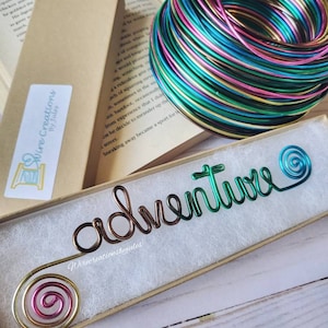 May include: A green, gold, pink, and blue wire bookmark with the word "adventure" written in wire. The bookmark is in a small box with a white interior.