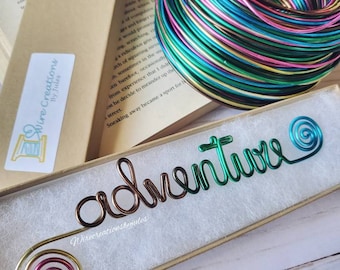 May include: A green, gold, pink, and blue wire bookmark with the word "adventure" written in wire. The bookmark is in a small box with a white interior.