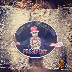 May include: Oval sticker featuring a cartoon image of Captain Spaulding in a patriotic Uncle Sam-style outfit. The sticker has the words "CAPTAIN SPAULDING" and "PRESIDENT" in a circular design. The sticker is on a rough, textured surface.