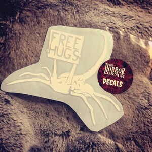 May include: White vinyl sticker of a monster holding a sign that says "Free Hugs". The sticker is on a gray fuzzy surface. The sticker is from "The Horror Corner Decals".