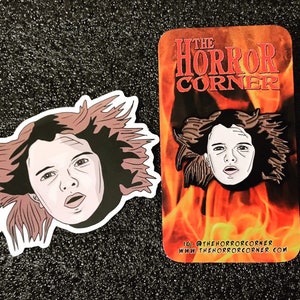 May include: A black and white enamel pin featuring a cartoon illustration of a woman with long, flowing hair. The pin is set against a red and orange background with the text "The Horror Corner" in white. The text "IG: @thehorrorcorner www.thehorrorcorner.com" is printed below the image.