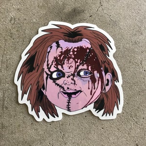 May include: A sticker featuring a cartoon illustration of a bloody, stitched-up doll's face with brown hair. The doll has a stitched-up mouth and a sinister smile.