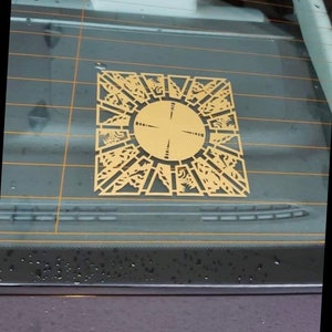 May include: A gold-colored sticker of a complex geometric design, resembling a puzzle or a maze, is adhered to a car window. The design features a central circle with radiating lines and intricate patterns.