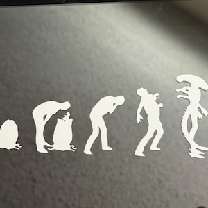 May include: A white vinyl sticker depicting the evolution of a human into an alien creature. The sticker shows a series of figures, starting with a human in a crouched position, then a human standing upright, then a human with a more alien-like head, and finally a fully formed alien creature with a long, thin tail.