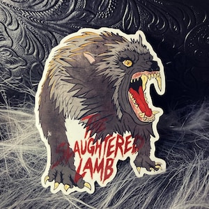 May include: A sticker featuring a snarling, gray and black werewolf with red eyes and sharp teeth. The werewolf is standing on its hind legs with its front paws raised. The text "The SLAUGHTERED LAMB" is written in red and black letters below the werewolf.