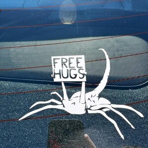 May include: A white car window sticker featuring a cartoon alien holding a sign that says "FREE HUGS".