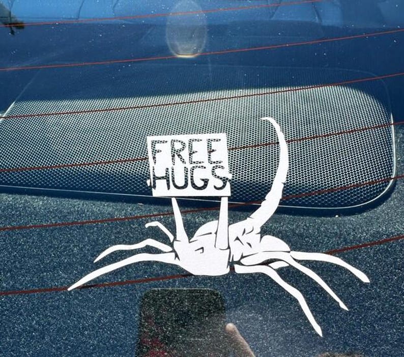 May include: A white car window sticker featuring a cartoon alien holding a sign that says "FREE HUGS".