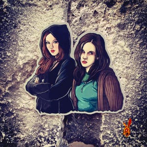 May include: A sticker featuring two women with stylized portraits. One woman wears a black hooded jacket, the other a teal top and brown cardigan. The sticker is placed on a textured, gray surface.