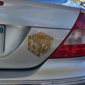 May include: A silver car with a gold sticker of a complex, intricate cube on the back bumper.