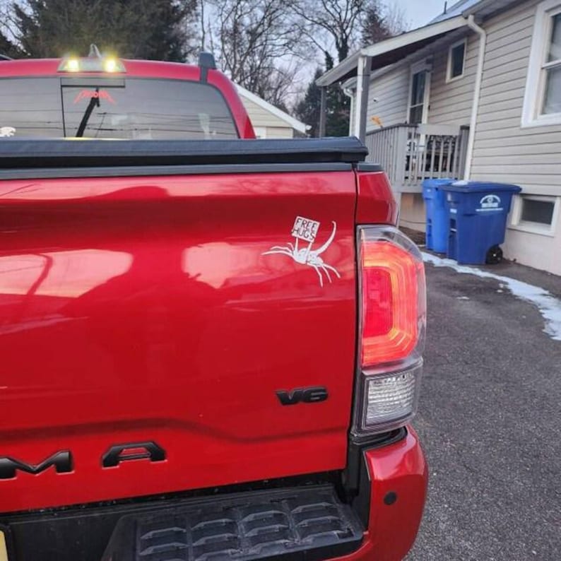 May include: A red pickup truck with a white spider sticker on the back. The sticker says "Free Hugs".