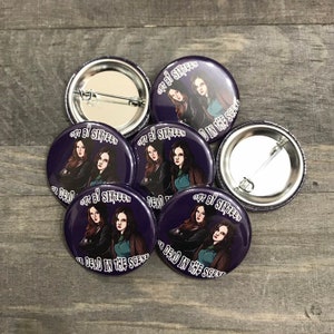 May include: A collection of purple button pins with a graphic of two women and the text "OUT BY SIXTEEN OR DEAD IN THE SCENE". The pins have a metal backing and a safety pin closure. The pins are arranged on a wooden surface.