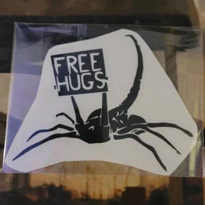 May include: Black and white sticker of a scorpion holding a sign that says "FREE HUGS".