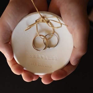 Puede incluir: Un plato de cerámica blanco con dos anillos de boda. El plato tiene los nombres "SEE ALEX" y la fecha "11 9 DEC 2015" grabados. Los anillos se sujetan con un lazo de cuerda.