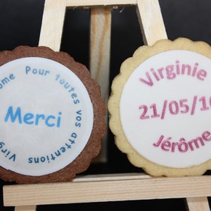 Peut inclure: Deux biscuits ronds avec glaçage blanc et bords festonnés. L'un est au chocolat avec le texte bleu "Merci". L'autre est à la vanille avec le texte rose "Virginie 21/05/16 Jérôme".